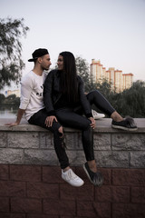 Fototapeta na wymiar vertical image beautiful young girl in black clothes gently looks into the eyes of a stylish young man sitting with his back on a concrete fence in a park on a background of tall buildings