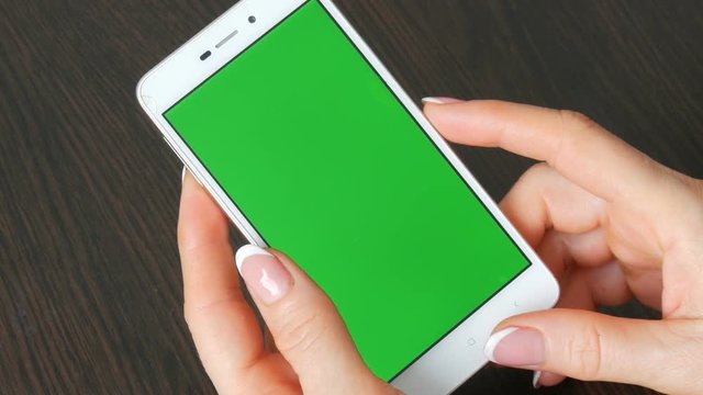 Female hands with beautiful French manicure take a white smartphone with Green Screen. Using Smartphone,Holding Smartphone with Green Screen on a stylish black wooden table