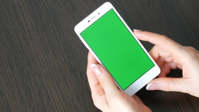 Female hands with beautiful French manicure take a white smartphone with Green Screen. Using Smartphone,Holding Smartphone with Green Screen on a stylish black wooden table