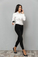 Full length image of pleased businesslike woman in formal wear walking and speaking on mobile phone with takeaway coffee in hand, isolated over gray background