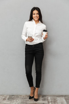 Full length image of attractive business woman in formal wear standing and holding mobile phone with paper cup of takeaway coffee in hand, isolated over gray background