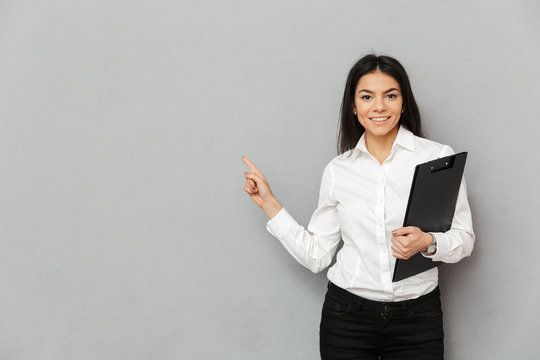 Portrait of office woman with long dark hair wearing white shirt smiling and pointing finger aside on copy space while holding folder with papers, isolated over gray background