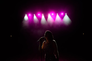 Girl in long gown performing on stage. Girl singing on the stage in front of the lights. Silhouette of singer standing on stage at microphone.