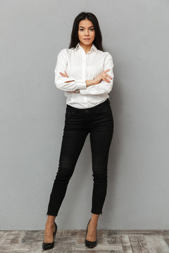 Full length image of caucasian woman with long brown hair wearing office clothing smiling at camera while posing with arms folded, isolated over gray background