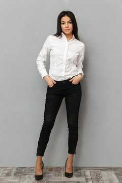 Full length image of attractive woman in white shirt and black trousers smiling at camera while posing with arms in pockets, isolated over gray background