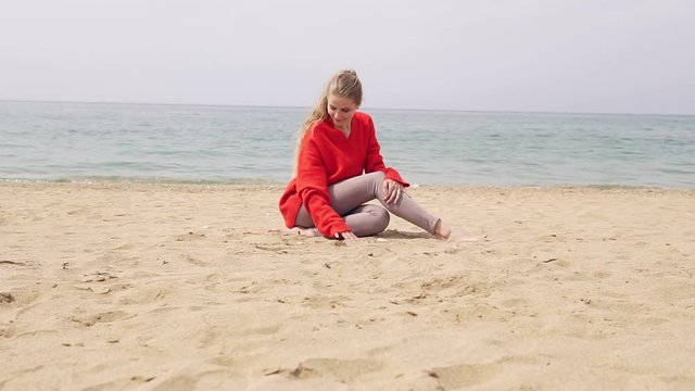 blonde girl is sitting on the sand by the sea and posing for a photo shoot
