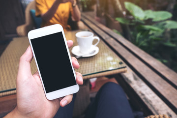 Mockup image of hand holding white mobile phone with blank black screen in cafe and blur woman in background