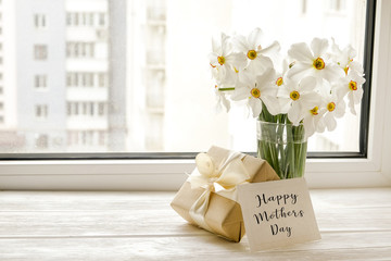 White yellow daffodil, narcissus flowers in glass vase on wooden windowsill, no window wiev. Happy mother's day text greeting card and craft paper prsent. Close up, copy space, still life, background.