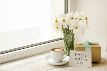 White yellow daffodil, narcissus flowers in glass vase on wooden windowsill, no window wiev. Happy mother's day text greeting card and craft paper prsent. Close up, copy space, still life, background.