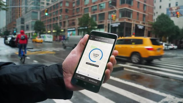 A man holds a smartphone running an ride sharing app in Manhattan on a rainy day. The screen reads Location Driver. Fictional interface.  	