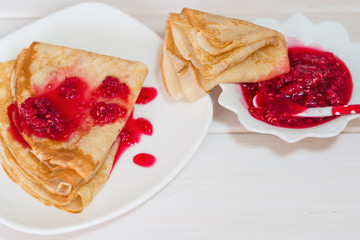 Pancakes with raspberry jam on a white plate stand on a natural wooden Board and a light wooden table 