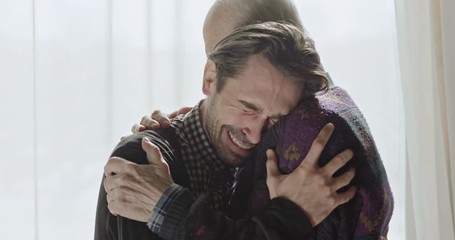 Side view of senior embracing adult man crying from happiness standing against window in sunlight.