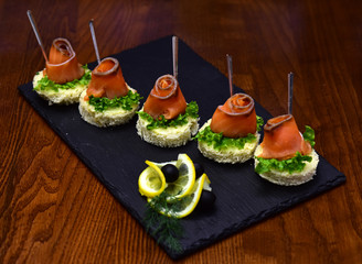Cold appetizers, canape with red fish, wooden background. Canape with salad leaves, red fish, lemon and dill. Delicious snacks served in restaurant on black dishes. Restaurant dish concept.