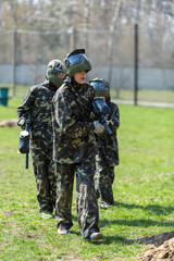 Boy in the camouflage holds a paintball gun  in one hand and protective helmet , standing on the field with group of players on the background