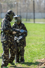 Fototapeta premium Boy in the camouflage holds a paintball gun in one hand and protective helmet , standing on the field with group of players on the background
