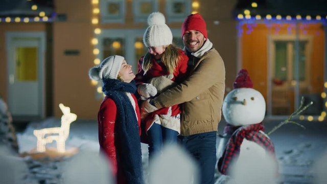Happy Young Family Portrait in the Falling Snow, Father Holds  Daughter and Embraces His Wife. Smiling Family Enjoys Winter Holiday in the Front Yard of their Idyllic House Decorated with Garlands.