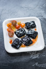 Black ravioli with cherry tomatoes and grated parmesan cheese served on a white plate, grey concrete background, vertical shot