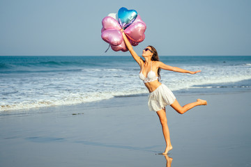 young and beautiful girl holding helium balloons air, running and jumping for joy at the sea....