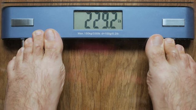 A severely overweight person weighing herself or himself on a digital scale. Man standing on weight scales with bare foot. Overeating diabetes.