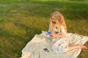 Beautiful charming barefoot long curly blonde hair teenage girl wearing a long light dress outdoors on a picnic paints with watercolor drawing sitting on a blanket on the grass.