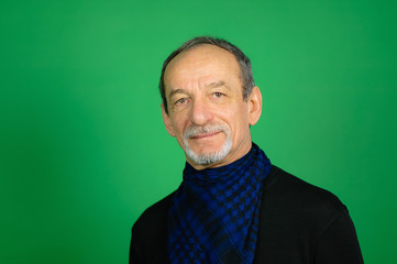 Fototapeta premium Gray-haired Senior Man with Well-trimmed Beard in Black Sweater and Blue Chequered Scarf Around his Neck Looking at the Camera on a Green Background in Studio