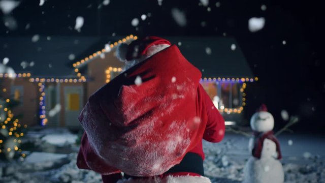 Authentic Santa Claus Carries Red Bag Over His Shoulder and Walks into Front Yard of the Idyllic House Decorated with Lights and Garlands. Magical New Year's Eve with Falling Snow.