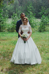 Fototapeta na wymiar Wedding couple is hugging in the green forest. Bride in satin dress with train and tulle veil is holding a bouquet and smiling. High green trees and bright emerald green grass outdoors. Warm embrace.