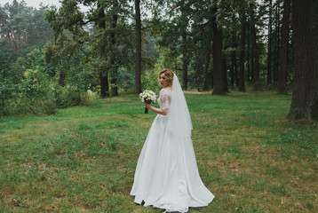 Bride in satin lace dress with train and tulle veil is holding a bouquet. High green trees and green grass outdoors. Smiling wedding bridal portrait in the wood. Eustoma and roses flowers in forest.