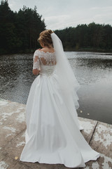 Bride in satin lace dress with train and tulle veil is standing on the lake bridge. High green trees and water in the forest. Romantic wedding portrait from the back.