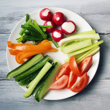 White plate with vegetables for a vegetarian salad. Radishes, tomatoes, celery, bell pepper, onion and cucumber. White wooden kitchen table. Close up. Top view. Toned photo.
