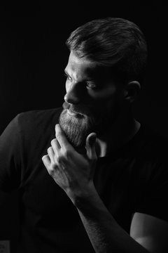 Black and white image of attractive pensive young man looks into the distance stroking his beard over black background