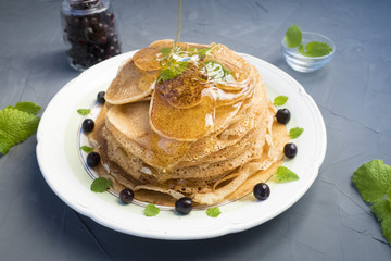 Pancakes are located in a plate on a gray background, decorated with berries and mint leaves. Maple syrup pours pancakes.