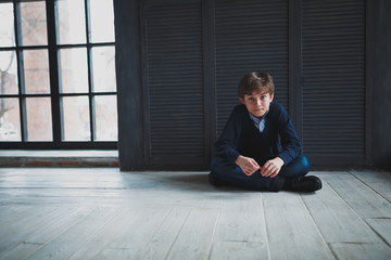 Sad teen boy in a blue shirt and jeans sitting on the floor near the dark blue walls in the room and looks in front of him. In the background large window.