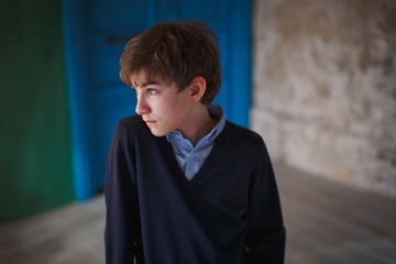 Serious pensive cute teen boy standing in loft style room and looking away. In the background there is a green wall and a bright blue door.