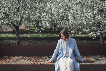 A young pretty brunette woman with short hair in bright romantic clothes and a blue coat sits on a bench in a blossoming park with cherry trees on a bright sunny day.