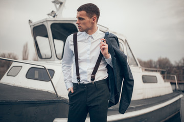 Young man with jacket in his hand stands on pier next to boat.
