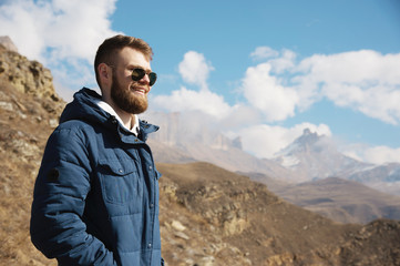 Hipster traveler in a down jacket and sunglasses stands on a mountain slope against the backdrop of epic rocks and smiles. The concept of finding happiness in the mountains