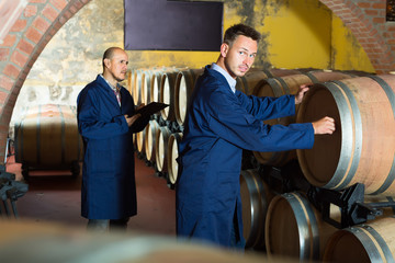 Fototapeta na wymiar two smiling men in uniforms taking notes in cellar with wine woods