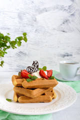 Whole wheat Belgian waffles with whipped cream, freshly chopped strawberries, mint leaves and chocolate on white wooden background. Homemade healthy breakfast.