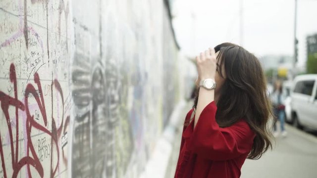 Beautiful young woman with dark hair wearing a red shirt is looking at the Berlin wall on a sunny summer day. Left to right pan real time medium shot