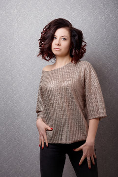 A brown-haired woman in a fashionable sweater and with a stylish hairstyle poses on the gray vintage wall.