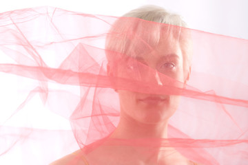 Young woman peering through shroud of pink chiffon fabric.