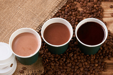 Three take away green paper cups with coffee beans and hot chocolate drink on wooden background, top view, close-up
