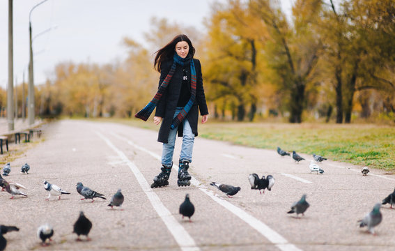A pretty girl in a coat and jeans with a long scarf happily skates in the autumn park with pigeons. Fighting the autumn depression.