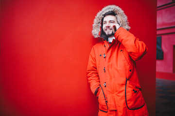 handsome young male student with a toothy smile and a beard stands on a red wall background in a bright red winter jacket with a hood with fur in winter. Uses a mobile phone, talk and call