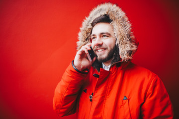 handsome young male student with a toothy smile and a beard stands on a red wall background in a bright red winter jacket with a hood with fur in winter. Uses a mobile phone, talk and call