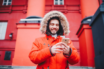 handsome young male student with toothy smile and beard stands on red wall background, facade of educational institution in red winter jacket with hood with fur, Uses finger on screen of mobile phone