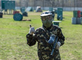 Boy in the camouflage holds a paintball gun  in one hand and protective helmet , standing on the field with group of players on the background