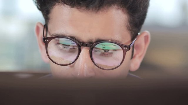close up of a young  busy Indian businessman wearing eyeglasses working on computer. Reflection of computer screen is seen in the eyeglasses of an handsome male entrepreneur in modern corporate office