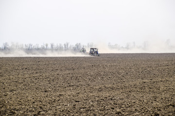 The tractor harrows the soil on the field and creates a cloud of dust behind it